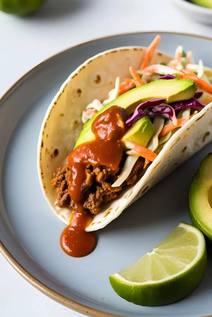 Overhead view of tacos filled with baked fish, slaw, avocado, and lime wedges on a plate.