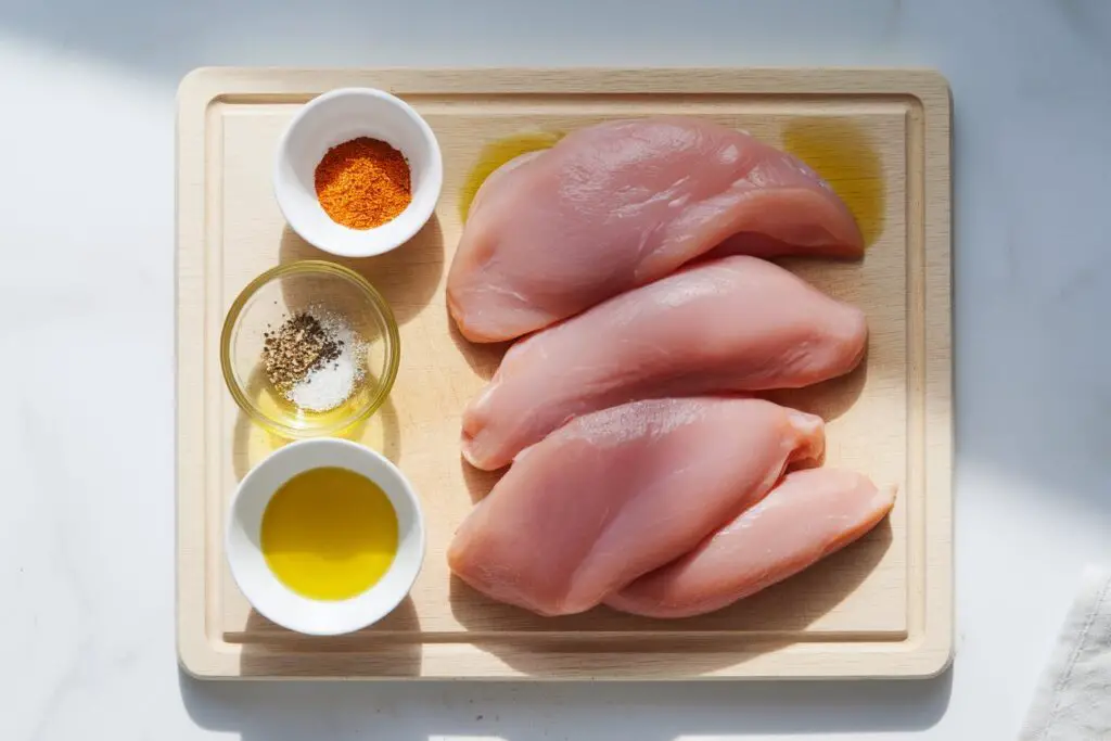 Raw boneless, skinless chicken breasts on a cutting board, seasoned with spices and olive oil.